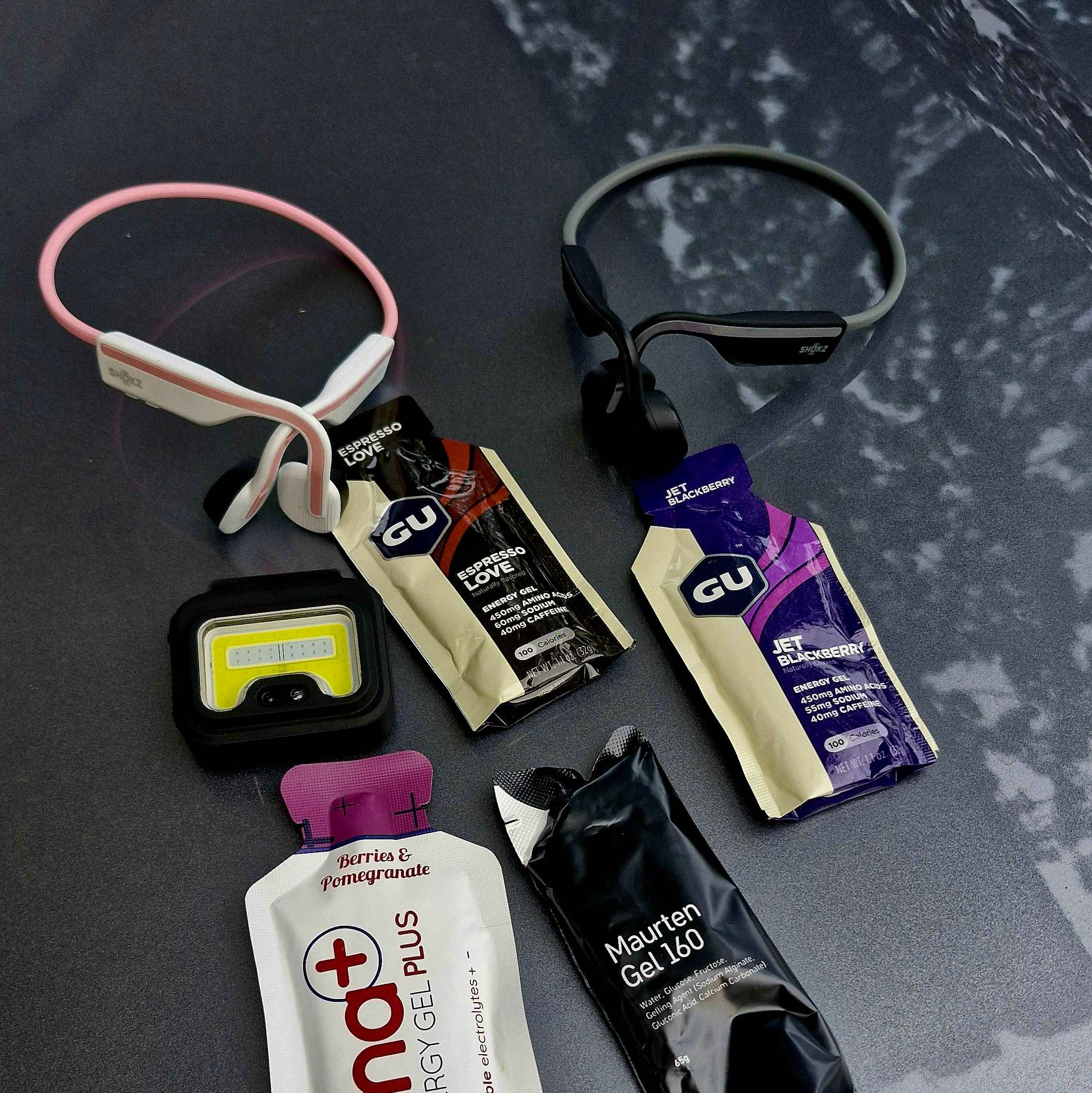 Top-down view of two pairs of Shokz bone conduction headphones (pink/white and black), four packs of assorted energy gels (GU Espresso Love, GU Jet Blackberry, Hüma Berries & Pomegranate, and Maurten Gel 160), and a small square LED safety clip-on light.